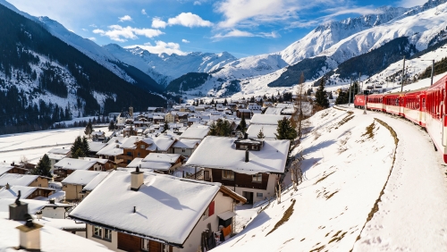 Glacier Express fährt durch eine verschneite Landschaft oberhalb eines sonnigen Dorfes~3µ3