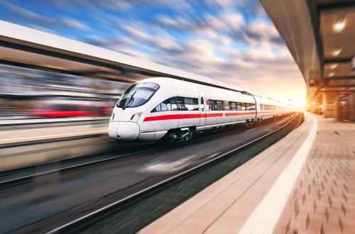 © den-belitsky - stock.adobe.com Weiße moderne Hochgeschwindigkeitszug in Bewegung auf Bahnhof bei Sonnenuntergang. Zug auf Bahnstrecke mit Bewegungsunschärfe in Europa abends. Bahnsteig. Industrielandschaft Bahntourismus